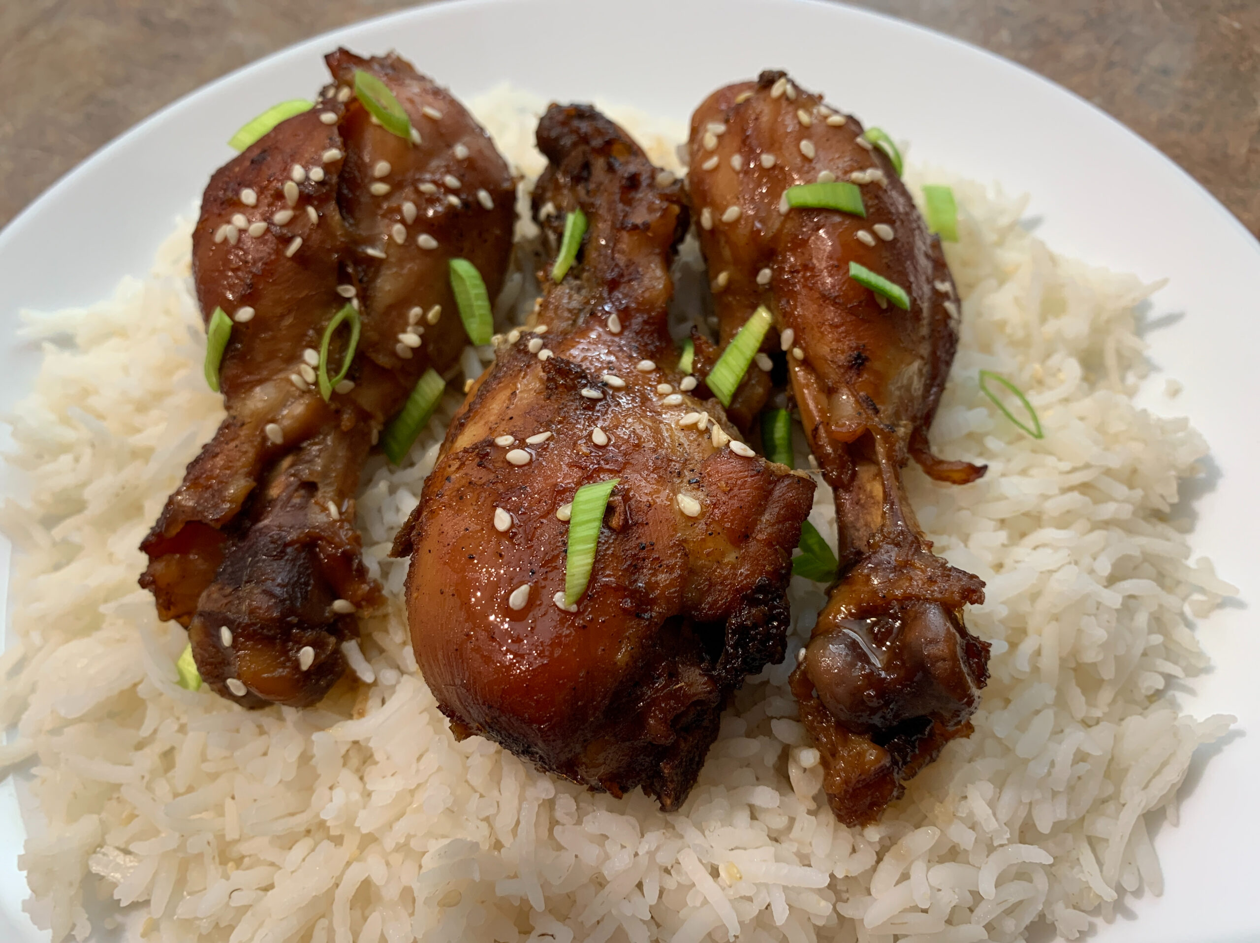 Plate of Asian baked chicken drumsticks glazed with a savory sauce, accompanied by steamed rice, garnished with green onions and sesame seeds.