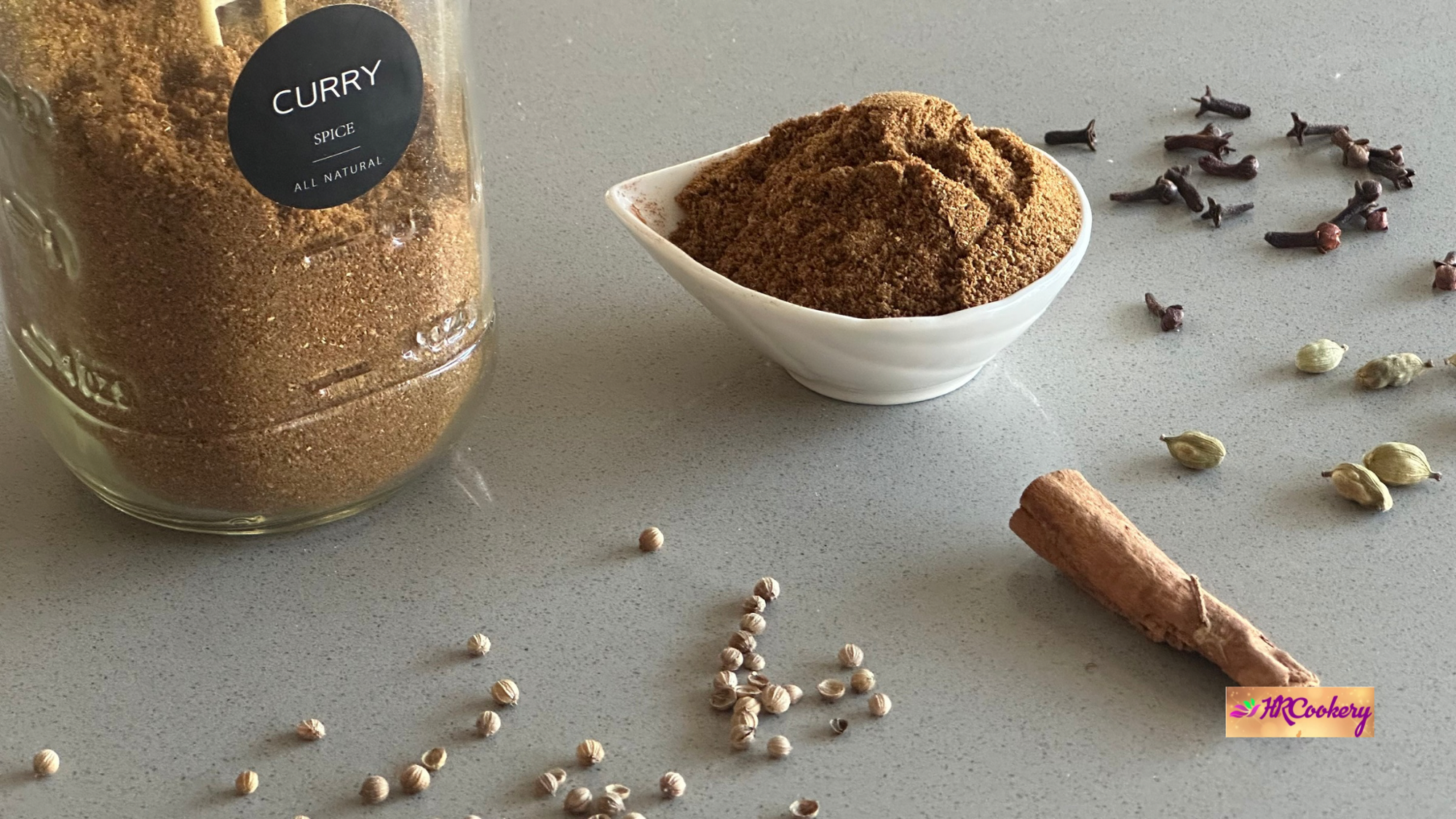 Homemade Ceylon curry powder in a white bowl with whole spices like cinnamon, cloves, cardamom, and coriander seeds on a kitchen counter.