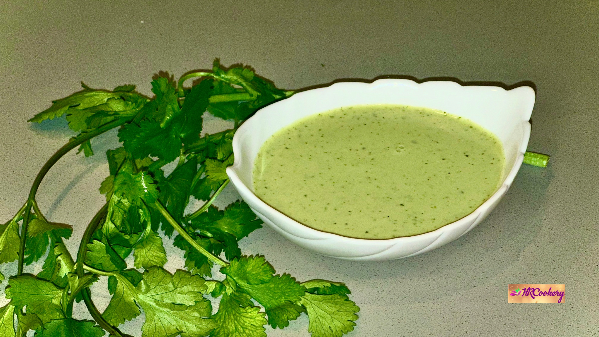 A white leaf-shaped bowl filled with creamy green cilantro garlic sauce, placed beside fresh cilantro on a light countertop.