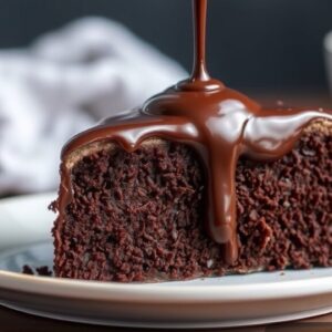 A rich chocolate mud cake slice on a white plate, garnished with a dollop of whipped cream and a sprinkle of cocoa powder, with a fork beside it.